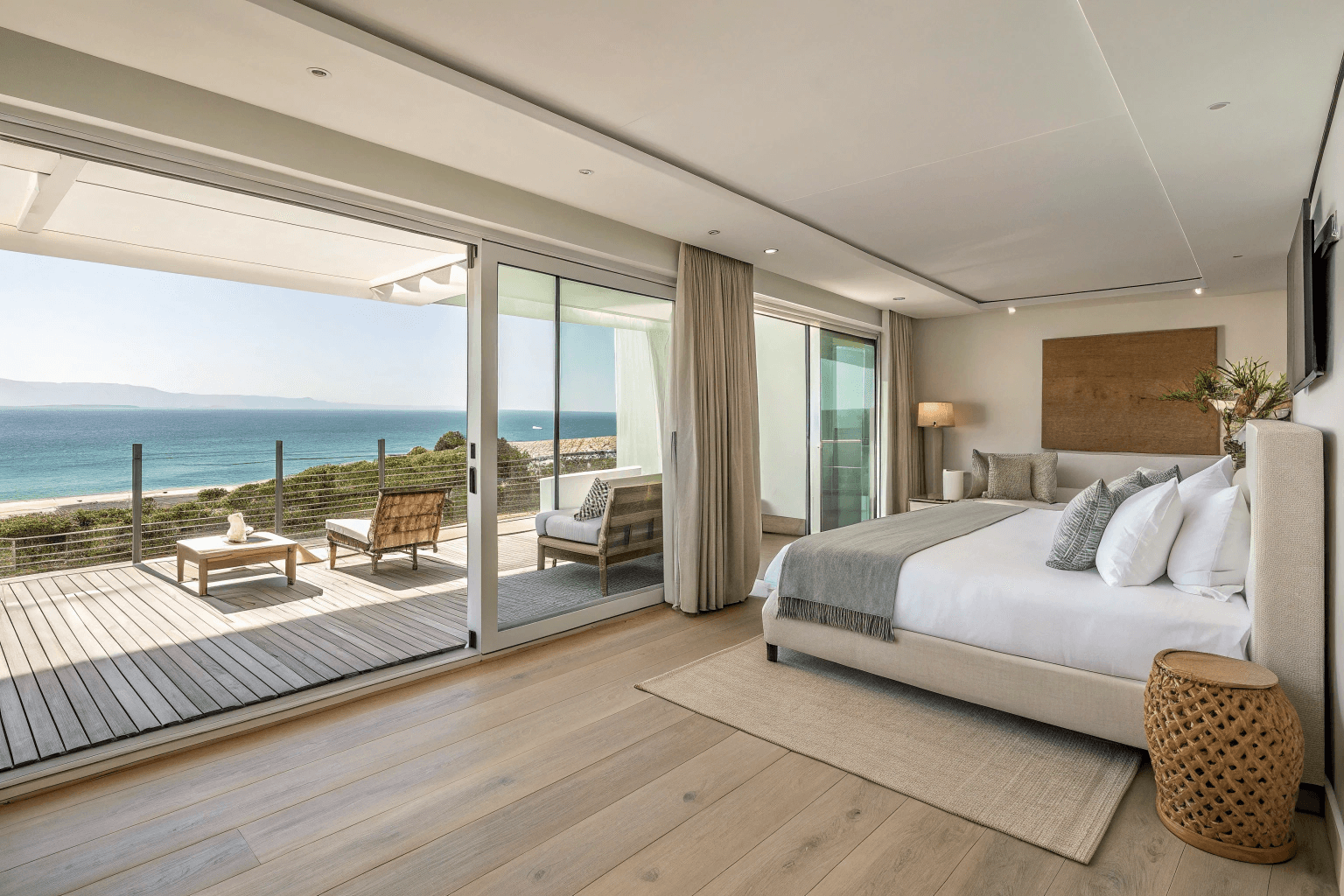 spacious open-plan suite with king bed, sofa lounge, light wood flooring, and ocean views through sliding glass doors.